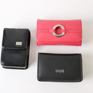 May include: Three leather accessories: a black case with a silver emblem, a coral-colored wallet with a silver ring detail, and a black cardholder with "IXUS" printed on it. All items are on a white background.