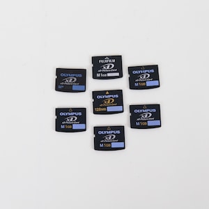May include: A collection of seven black xD-Picture Cards arranged in a cluster. Six cards are branded Olympus with capacities of 1GB or 128MB, and one card is branded Fujifilm with a 1GB capacity. Each card features white and blue text labels.