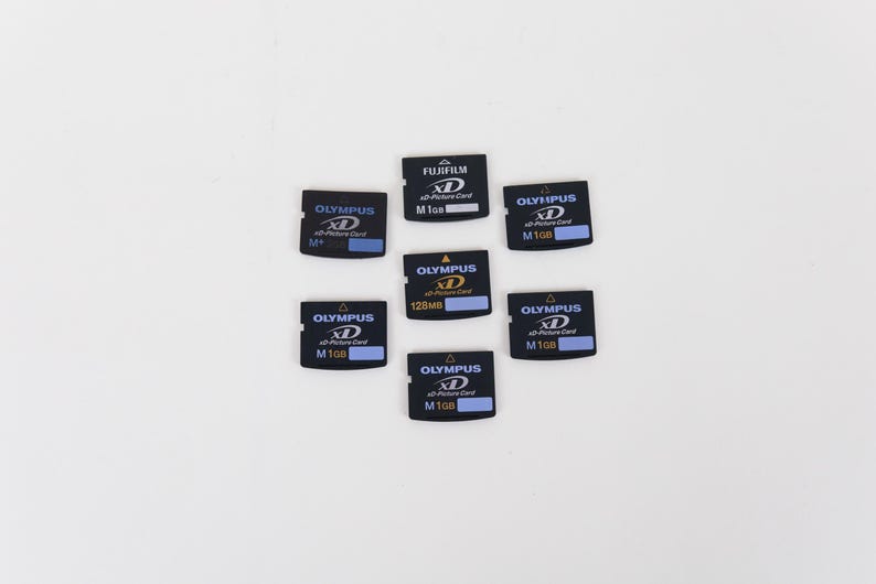 May include: A collection of seven black xD-Picture Cards arranged in a cluster. Six cards are branded Olympus with capacities of 1GB or 128MB, and one card is branded Fujifilm with a 1GB capacity. Each card features white and blue text labels.