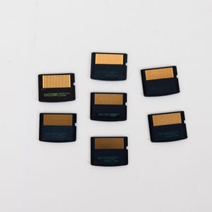 May include: Six black rectangular SD memory cards arranged in a cluster. Each card features a gold-colored contact strip along the top edge and printed text in green on the lower portion of the black plastic casing, showing storage capacity.
