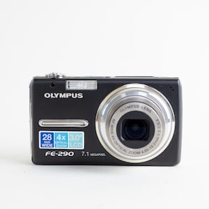May include: A black Olympus FE-290 digital camera featuring a silver lens ring, a built-in flash, and a front-facing label displaying 28mm wide, 4x optical zoom, and 3.0 inch LCD specifications. The camera body is rectangular with rounded edges.