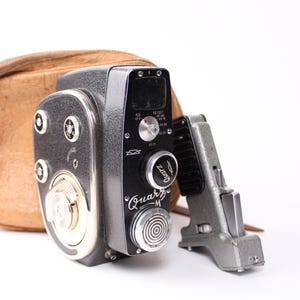 Puede incluir: Una cámara de cine vintage negra y plateada con una funda de cuero marrón. La cámara tiene una lente plateada y un dial negro con el texto "Quartz-M" y "Quartz" en él.