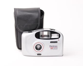 Pode incluir: Uma câmera de filme de 35 mm Hanimex VC3200 prateada com flash embutido e uma capa de couro preta.