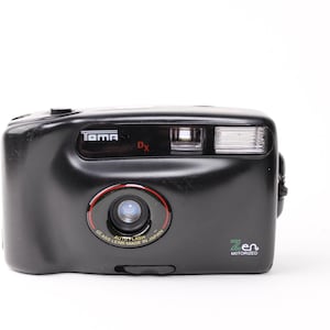 May include: A black Tama Zen DX autofocus camera with a glass lens made in Japan. The camera has a built-in flash and is motorized.
