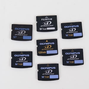 Puede incluir: Una colección de siete tarjetas de memoria xD-Picture negras de Olympus y Fujifilm. Cada tarjeta muestra logotipos de marca, el texto xD-Picture Card y capacidades de almacenamiento de 1 GB, 2 GB y 128 MB, con pequeñas etiquetas azul claro.