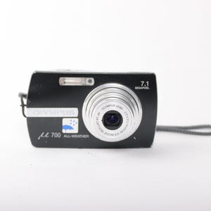 May include: A black Olympus digital camera with a silver lens, featuring the text 7.1 MEGAPIXEL, OLYMPUS, and 700 ALL-WEATHER with a blue umbrella icon. The lens is labeled OLYMPUS LENS AF 3X OPTICAL ZOOM 6.5-19.5mm 1:3.4-5.7. Includes a grey wrist strap.