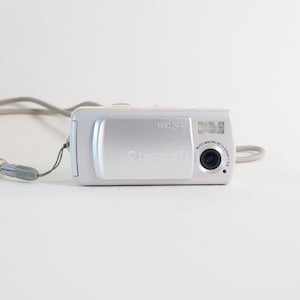 May include: A silver Sony Cyber-shot digital camera with a sliding lens cover, a built-in flash, and a circular lens with text reading Auto Macro AF f=5.3mm 1:2.8. The camera includes a light gray wrist strap with a clear plastic bead.