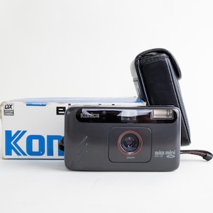 以下が含まれることがあります： ダークグレーのKonica Big Mini BM-20 Jr 34mmフィルムカメラ。黒いレザー製のキャリングケースと、オリジナルの白と青の紙箱が付属しています。カメラには赤いリングが付いたフロントレンズ、内蔵フラッシュ、リストストラップが備わっています。