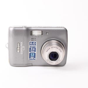 May include: A silver Nikon Coolpix L2 digital camera featuring a 6.0 Megapixels label, 3x Zoom, and Face-priority AF text on the front. The lens is a Nikkor 3x Optical Zoom 6.3-19.2mm 1:3.2-5.3. The camera body has a built-in flash unit.