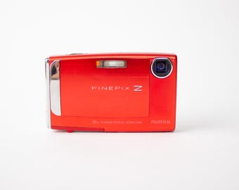 May include: A red Fujifilm FinePix Z digital camera. The camera has a silver accent on the side and a lens on the front. The text "FINEPIX Z" is printed on the front of the camera.