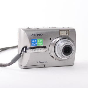 May include: A silver Olympus FE-140 digital camera featuring a 6.0 megapixel sensor and a 2.5 inch LCD screen. The front of the device displays the model name FE-140, the Olympus brand logo, and a gray wrist strap attached to the side of the body.