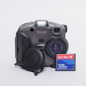 May include: A grey Kodak digital camera with a lens cap and a 128MB CompactFlash memory card. The camera has a built-in flash and the Kodak logo. The memory card is blue and red with the SanDisk logo.