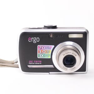 May include: A black and silver Ergo DC 7375 digital camera with a wrist strap. The front features a lens with 3X Super Zoom, a flash, and a sticker displaying 7.0 Mega Pixels, 3.0 Inch LCD, and 3X Optical Zoom specifications on the camera body.