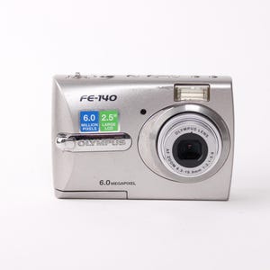 May include: A silver Olympus FE-140 digital camera featuring a 6.0 megapixel sensor and a 2.5-inch LCD screen. The front of the camera displays the model name FE-140, the Olympus brand logo, and lens specifications around the circular lens housing.