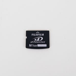 May include: A black Fujifilm xD-Picture Card memory card with white text. The card features the Fujifilm logo, the xD logo, the text xD-Picture Card, and the capacity label M 1GB with a small white rectangular space for writing.