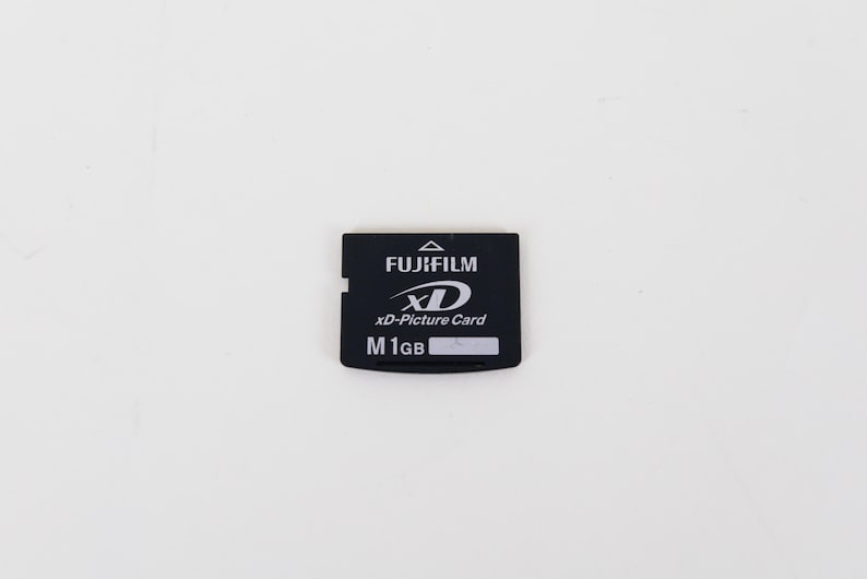 May include: A black Fujifilm xD-Picture Card memory card with white text. The card features the Fujifilm logo, the xD logo, the text xD-Picture Card, and the capacity label M 1GB with a small white rectangular space for writing.