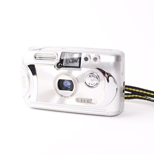 May include: Silver point-and-shoot film camera with a black and yellow wrist strap. The camera has a lens, flash, and various buttons. The body is shiny and reflective, with a clean, modern design.
