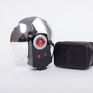 May include: A vintage chrome camera flash with a black control panel featuring a red and blue dial. A black leather case with the text "Lucky Light" is next to the flash. The flash has a reflective, rounded head.