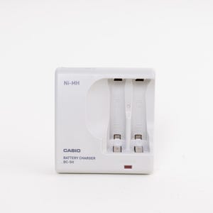May include: A white Casio battery charger, model BC-5H, with two charging slots. The charger has the text "Ni-MH" and a red indicator light. The device is designed to charge batteries.
