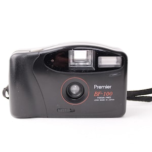 May include: A black Premier BF-100 focus free camera with a lens made in Japan. The camera has a built-in flash and a strap.