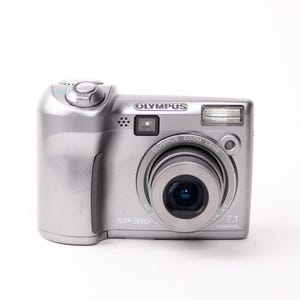 May include: Silver Olympus SP-310 digital camera. The camera has a zoom lens and a built-in flash. The camera has the text "OLYMPUS" and "SP-310" printed on it. The camera has a 7.1 megapixel resolution.