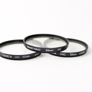 May include: Three black camera lens filters with UV protection. The filters are 55mm in diameter and have the brand names Tamica, Quantaray, and HMC printed on them.