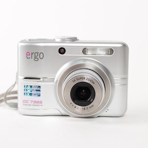 May include: A silver Ergo DC 7365 digital camera featuring a 3X super zoom lens with a focal length of 6.1-18.3 mm. The front of the camera displays the brand name, a flash unit, and technical specifications including 7.0 mega pixels and a 2.36 inch LCD monitor.