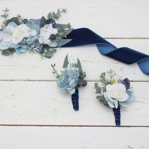 Pu&ograve; includere: Un set di tre boutonni&egrave;res floreali blu e bianche con nastro di velluto blu navy. Le boutonni&egrave;res sono realizzate con fiori di seta e verde.