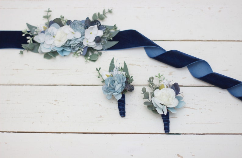 Pu&ograve; includere: Un set di tre boutonni&egrave;res floreali blu e bianche con nastro di velluto blu navy. Le boutonni&egrave;res sono realizzate con fiori di seta e verde.