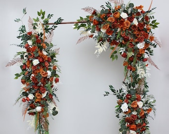 May include: A copper metal archway decorated with a cascading arrangement of artificial flowers. The flowers are a mix of orange, white, and green. The archway is standing on a wooden floor.