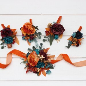 May include: Five small floral arrangements with orange, burgundy, and teal flowers. The arrangements are tied with a long orange ribbon.