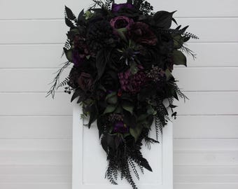 May include: A cascading floral bouquet featuring deep black and dark purple roses, peonies, and various foliage. The arrangement includes textured greenery and spiky thistle accents, creating a dense, layered composition of dark botanical elements.