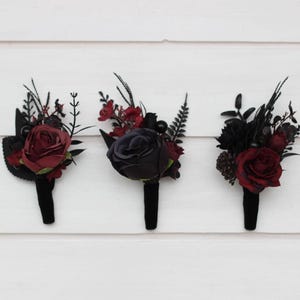 May include: Three black and burgundy floral boutonnieres with a single rose in each. The roses are burgundy, black, and dark red.