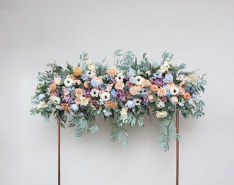 Op de afbeelding: Een bloemenboog gemaakt van kunstbloemen in tinten blauw, roze en perzik. De boog is gemaakt van verschillende bloemen, waaronder rozen, hortensia's en pioenen. De boog wordt ondersteund door twee gouden metalen palen.