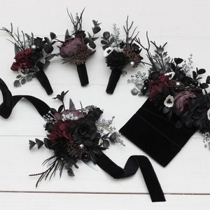 May include: A set of six black and burgundy floral buttonholes with silver accents. The buttonholes are arranged on a white surface with a black velvet pouch and ribbon.