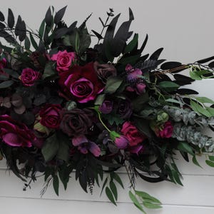 May include: A dark green and burgundy floral arrangement with deep red roses, burgundy foliage, and eucalyptus sprigs.