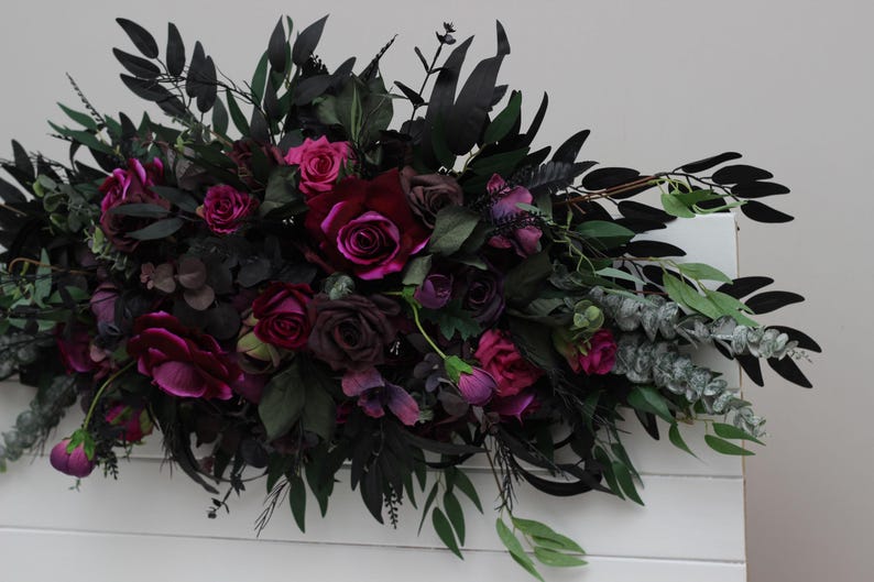 May include: A dark green and burgundy floral arrangement with deep red roses, burgundy foliage, and eucalyptus sprigs.