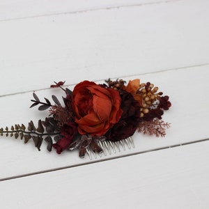 May include: A burgundy and orange floral hair comb with a large orange flower and smaller burgundy flowers. The comb is made of metal and has a decorative design.