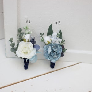 Pu&ograve; includere: Due boutonni&egrave;re con fiori bianchi e blu, verde e nastro blu scuro. Le boutonni&egrave;re sono etichettate con #1 e #2.