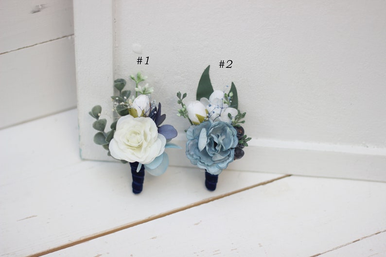Pu&ograve; includere: Due boutonni&egrave;re con fiori bianchi e blu, verde e nastro blu scuro. Le boutonni&egrave;re sono etichettate con #1 e #2.