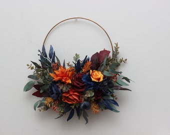 May include: A gold metal hoop wreath decorated with a cluster of faux flowers in shades of orange, red, and blue. The flowers are arranged in a cascading style, creating a full and lush look.