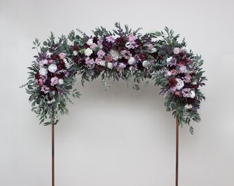May include: A floral arch arrangement featuring deep burgundy, soft pink, and white flowers mixed with various green eucalyptus leaves. The floral display is mounted on two thin, vertical copper-colored metal poles, creating a semi-circular shape.