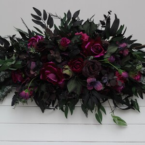 May include: A dark green and burgundy floral arrangement with deep red roses and greenery. The arrangement is on a white wooden background.
