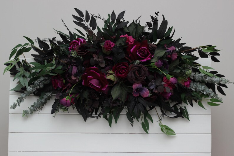May include: A dark green and burgundy floral arrangement with deep red roses and greenery. The arrangement is on a white wooden background.