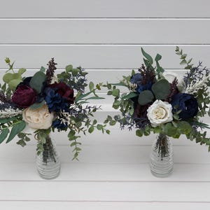 May include: Two bouquets of artificial flowers in clear glass vases. The bouquets feature burgundy, navy, and cream-coloured flowers, with green eucalyptus leaves and sprigs of white filler flowers. The vases are placed on a white slatted surface.