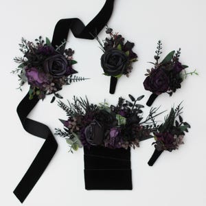 May include: A collection of dark floral accessories featuring deep purple and black artificial roses, dark foliage, and sprigs. The set includes a large floral arrangement with a black velvet ribbon, three smaller boutonnieres, and a floral hair piece.
