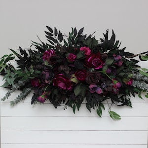 May include: A white wooden backdrop decorated with a dark green and burgundy floral arrangement. The arrangement features a variety of flowers, including roses, and greenery.
