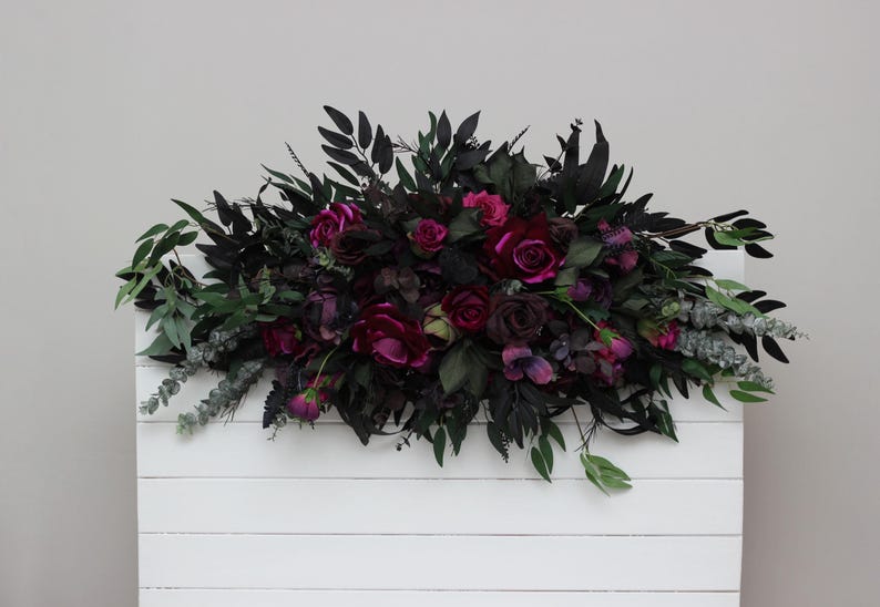May include: A white wooden backdrop decorated with a dark green and burgundy floral arrangement. The arrangement features a variety of flowers, including roses, and greenery.