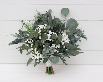 May include: A bouquet of greenery and white flowers, including eucalyptus and baby's breath. The bouquet is tied with a white ribbon and has a natural, rustic appearance. The leaves are various shades of green.