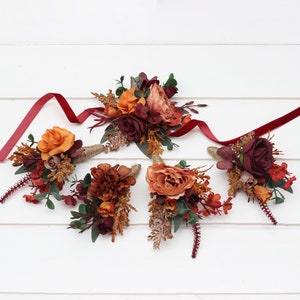May include: Five burgundy and orange floral buttonholes with red velvet ribbon. The buttonholes are arranged on a white wooden surface.